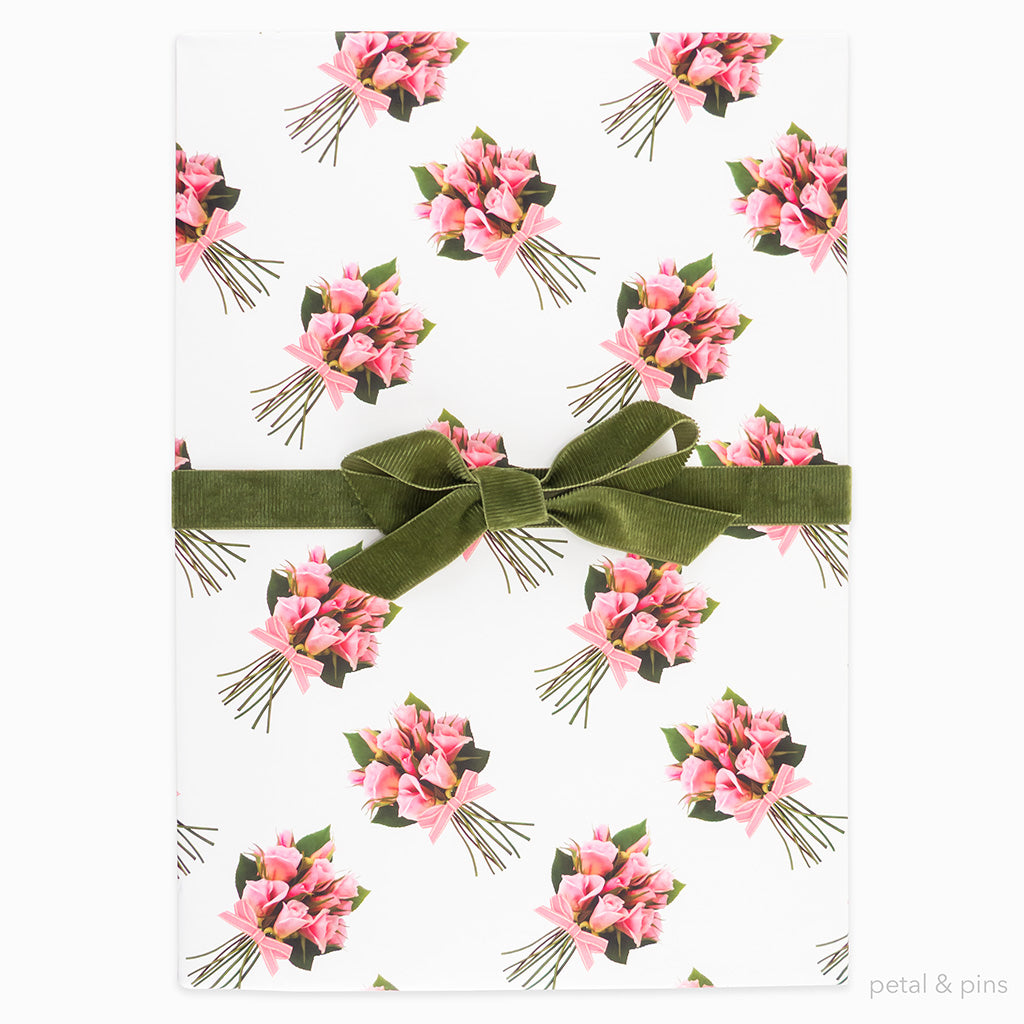 Rose posies wrapping paper by petal & pins. Floral patterned paper with pink flowers and green leaves on a white background, folded and tied with a green bow..