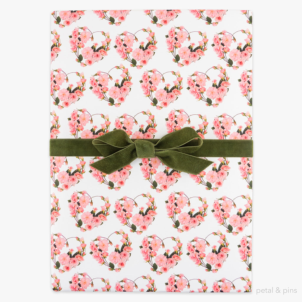 Rose hearts wrapping paper by petal & pins. Floral patterned paper with pink flowers and green leaves on a white background, folded and tied with a green bow.