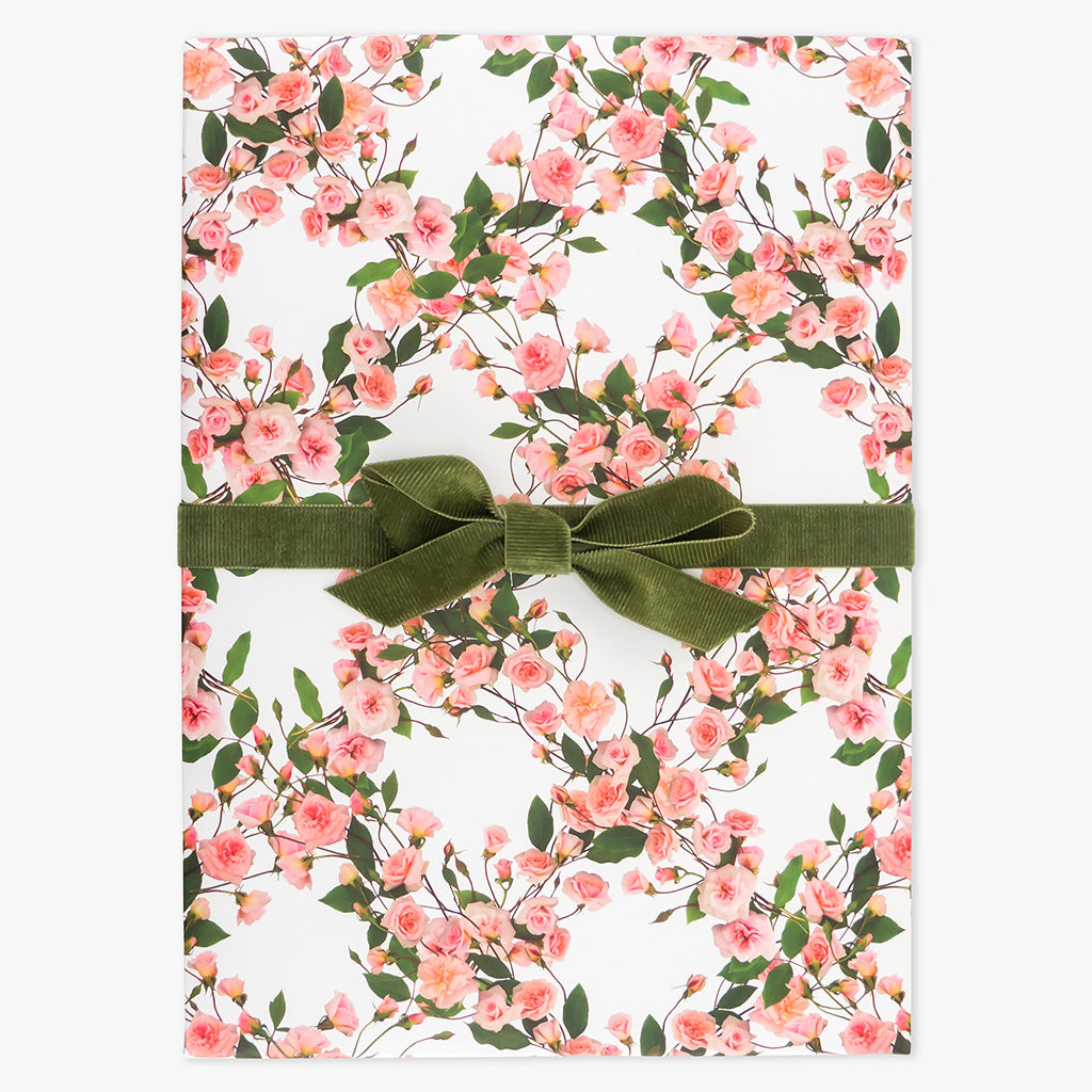 Rose posies wrapping paper by petal & pins. Floral patterned paper with pink flowers and green leaves on a white background, folded and tied with a green bow.